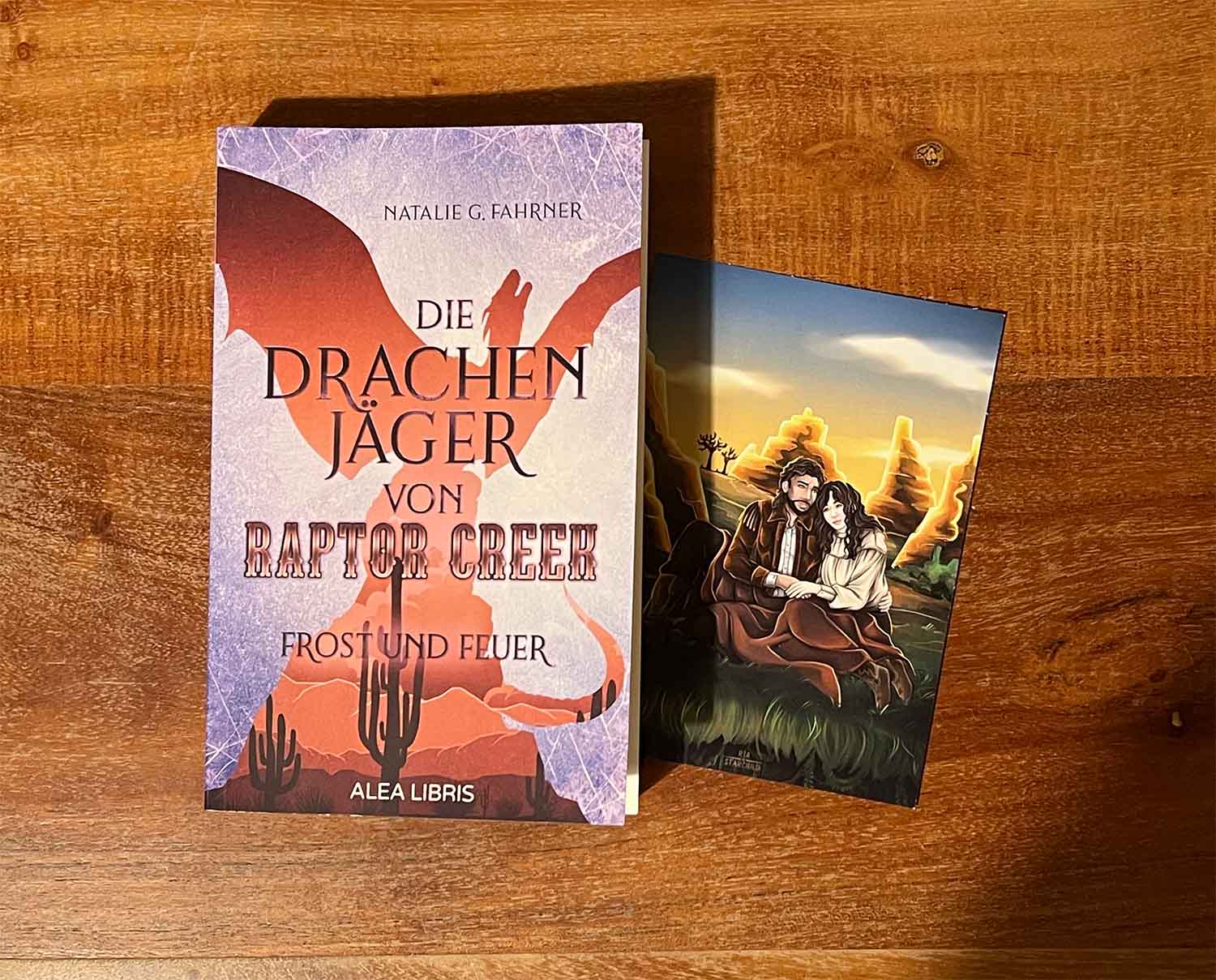 Das Bild zeigt das Buch "Die Drachenjäger von Raptor Creek" und eine Charakterkarte, auf der ein Paar in einer Canyon-Landschaft zu sehen ist.